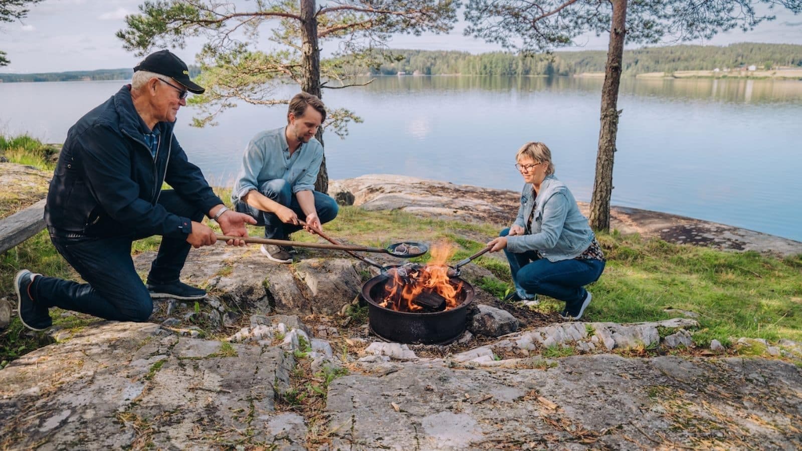1 dame og 2 menn som lager mat over et bål, med en sjø i bakgrunnen