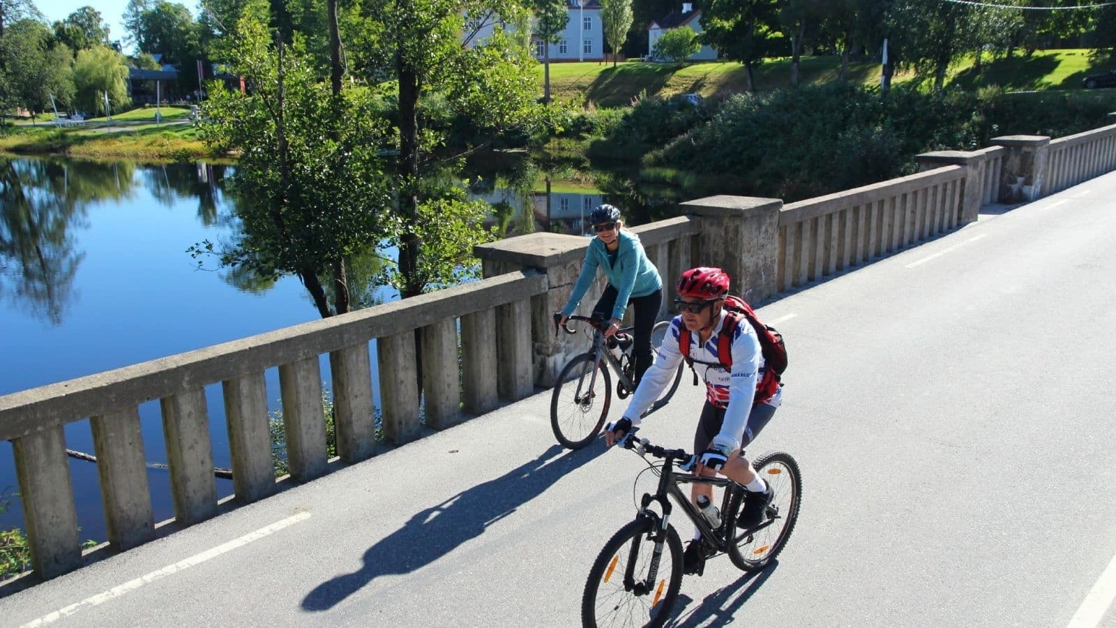 o syklister på en bro over vann omgitt av trær og hus i grønt landskap på Romerike